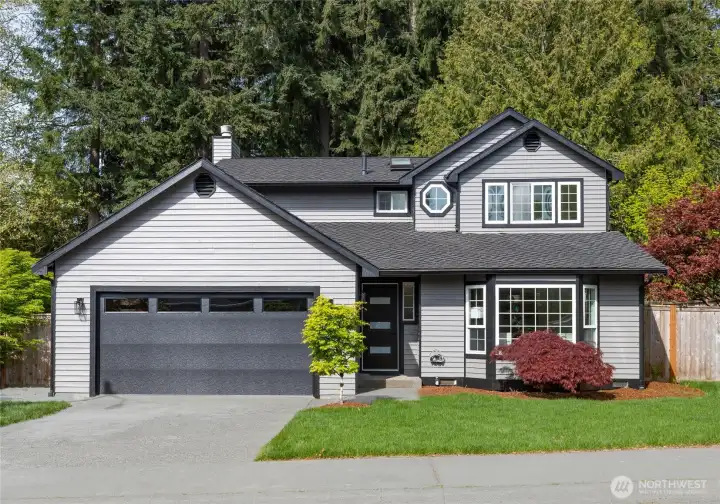 Charming two-story home with gray lap siding, bold black trim, and a statement garage door, set against a lush evergreen backdrop with mature Japanese maples and fresh bark landscaping.