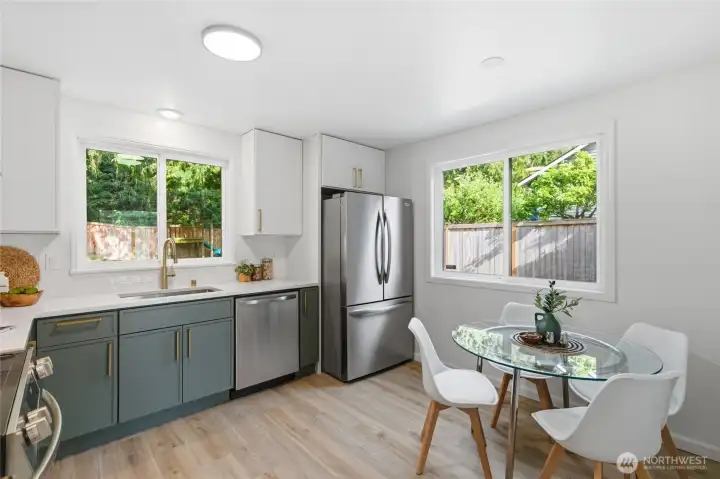 Updated kitchen with sage green lower cabinets, white uppers, quartz countertops, gold hardware, stainless appliances, and a breakfast nook — surrounded by windows with backyard views on two sides.
