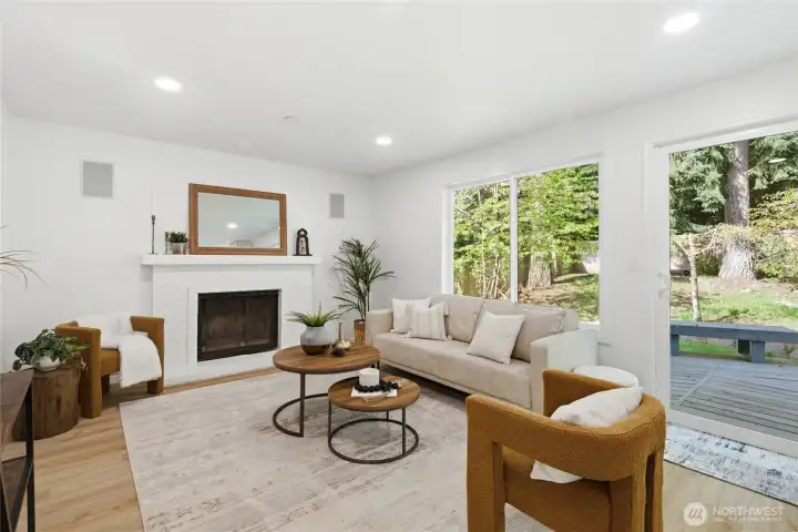 Cozy family room featuring a white brick fireplace, recessed lighting, in-wall speakers, and a sliding glass door to the deck with lush green views beyond.