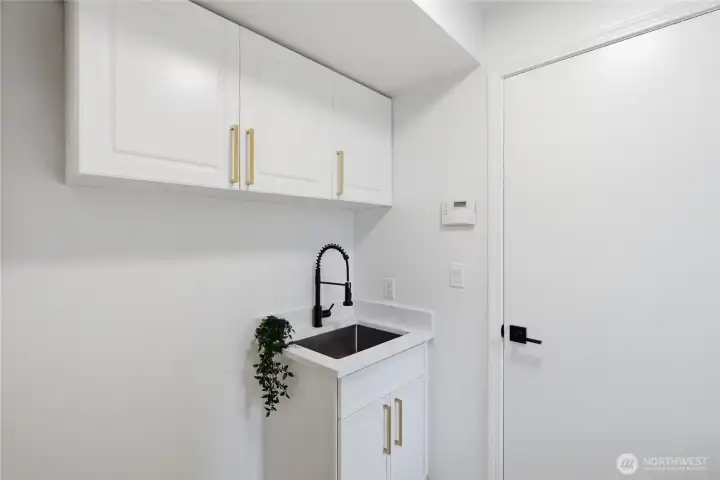 A well-appointed utility space featuring white cabinetry with gold hardware, a deep stainless sink with matte black faucet, and quartz countertop — functional and polished.