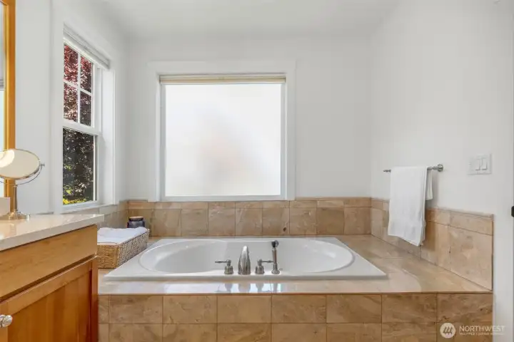 The soaking tub is surrounded by travertine tile and has obscured glass windows.