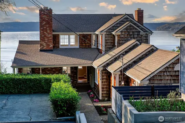 With its weathered cedar shingles, classic brick chimneys, and west-facing silhouette, this 1964 Cape Cod crowns a private quarter-acre hillside overlooking Puget Sound. From the moment you arrive, a sense of retreat takes hold—framed by hedge-lined terraces, generous parking, and a tree-canopied entry that hints at the home’s sweeping Olympic Mountain and Vashon Island views. Tucked within the sought-after Browns Point Improvement Club district, this is where coastal serenity meets timeless Northwest architecture.