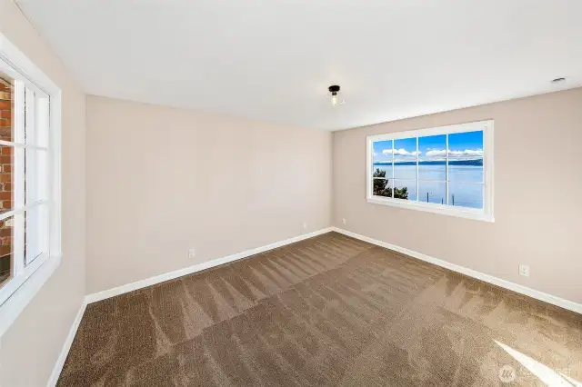 Wake each morning to panoramic views of Puget Sound and the Olympic Mountains beyond. This spacious bedroom, wrapped in light from dual exposures, offers a serene vantage point above Caledonia Cove—an ideal setting for rest, reflection, or creative retreat in the heart of Browns Point.