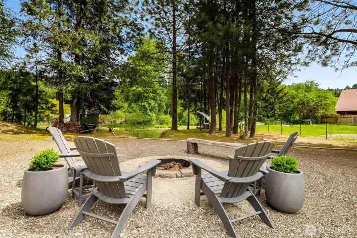 Outdoor fire pit area with gravel and surrounding seating under mature trees
