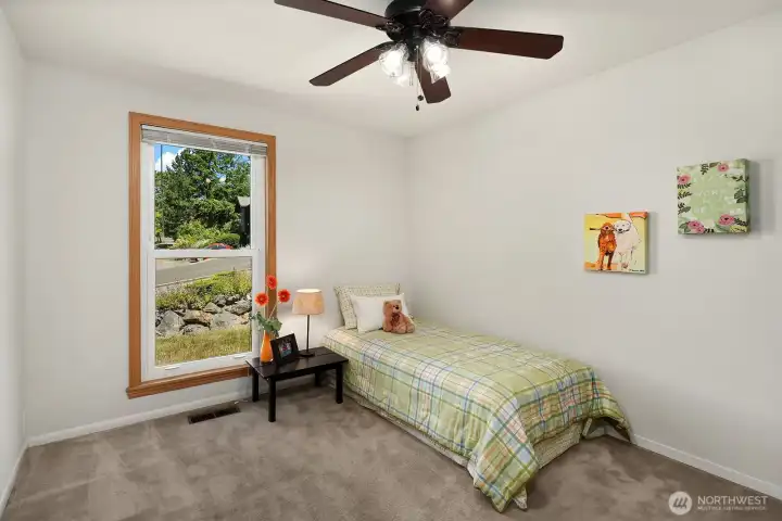 A bright, comfortable bedroom with soft carpeting, a ceiling fan, and a tall triple-pane window overlooking the landscaped front yard.