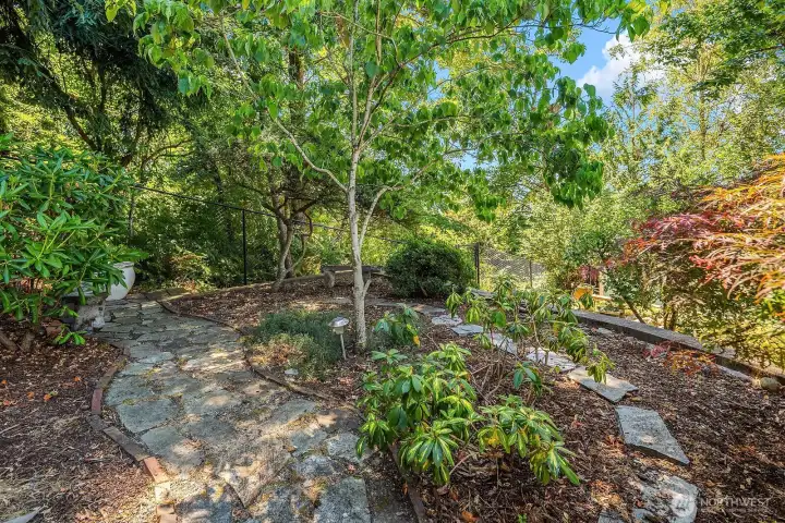 Paved pathways wind through the fully fenced backyard, connecting layered garden beds, mature landscaping, and the peaceful greenbelt beyond.