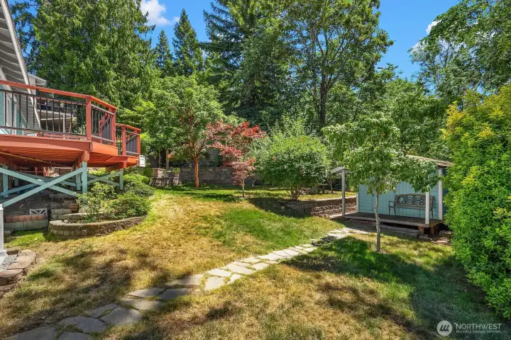 A paved garden path leads to the detached shed, surrounded by fruit trees, colorful maples, mature landscaping, and a fully fenced yard backing to greenbelt.