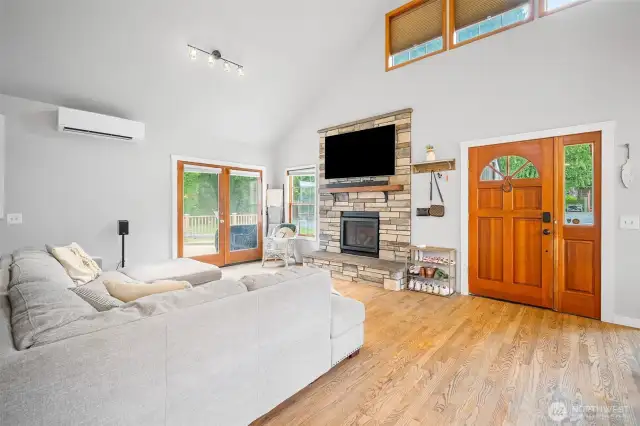 Stylish and welcoming living room featuring a vaulted ceiling, floor-to-ceiling stone fireplace, rich wood front door, and French doors that open to the spacious outdoor deck.