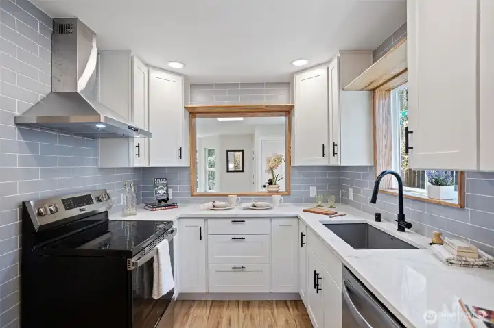 A kitchen reborn — new cabinetry, quartz countertops, blue-grey subway tile, stainless appliances, wall-mount range hood, and a wood-framed serve-through back to the living room.