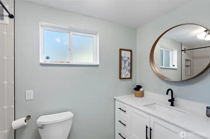 The full bath serves both daylight bedrooms — white shaker vanity, matte black hardware, warm wood-framed mirror, and a custom tile shower surround. Renovated, windowed, and finished with care.