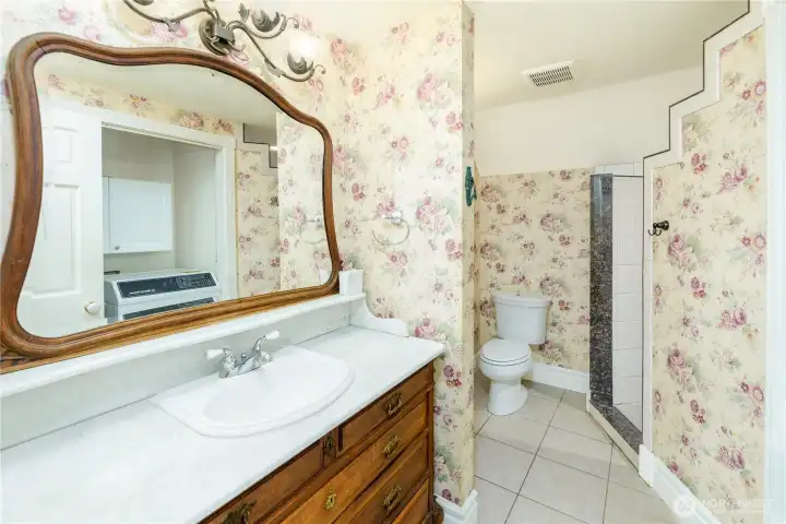 Charming guest bath featuring a vintage-inspired wood vanity with marble countertop, decorative floral wallpaper, and a walk-in tiled shower. Thoughtful architectural details and classic finishes create a warm, cottage-inspired retreat with timeless character. Charming guest bath featuring a vintage-inspired wood vanity with marble countertop, decorative floral wallpaper, and a walk-in tiled shower. Thoughtful architectural details and classic finishes create a warm, cottage-inspired retreat with timeless character.