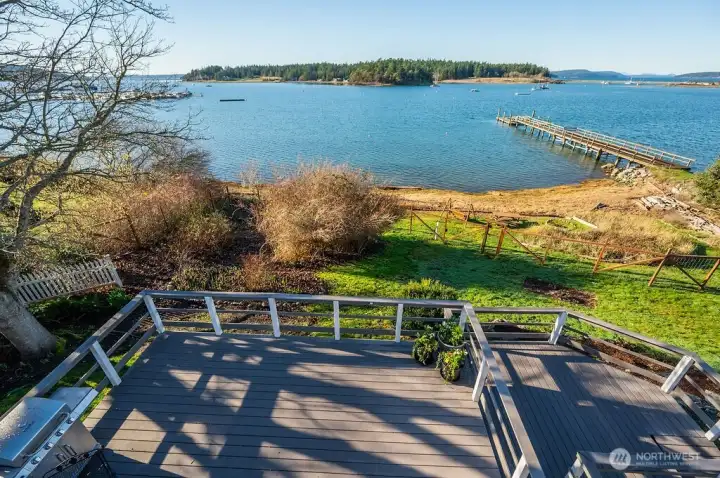 View across Fisherman Bay from main house, upper level deck (observation deck), looking down on main level deck, fenced garden, and then fire pit and picnic area on the beach. The peninsula and Spit Preserve (Lopez Island) seen across the water, with the entrance to Fisherman Bay off to the right