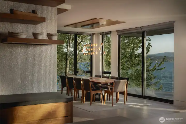 Positioned within a glass-wrapped corner, the dining area draws in the landscape and water beyond.