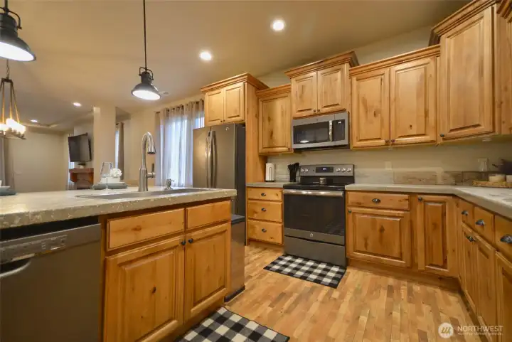 Beautiful open-concept kitchen featuring custom alder wood cabinetry and sealed builder-grade concrete countertops. Offers ample cabinet and drawer storage, a walk-in pantry, and a spacious island with breakfast bar. Designer lighting. Equipped with stainless steel appliances and distressed LVP flooring. Seamlessly flows into the dining and living areas, creating an ideal space for entertaining.