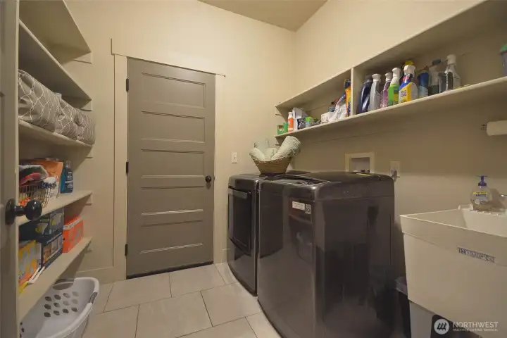 Functional laundry room with direct access to the spacious garage. Features tile flooring, a utility sink, and abundant storage with shelving above and along the opposite wall. Washer and dryer are negotiable.