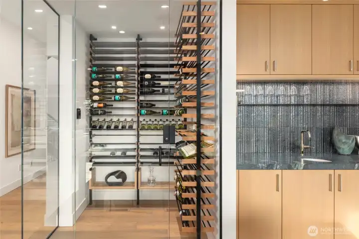 Sleek and contemporary temperature controlled wine room with custom designed shelving creates a light filled and artistic corner wall and flanks the dedicated dining space alongside wet bar featuring custom cabinets, desinger slab counters and tile backsplash, and hidden wood panelled Sub Zero Ice Maker