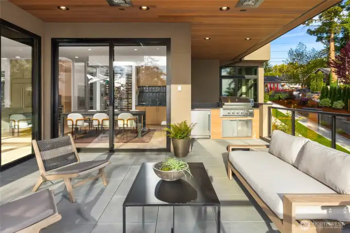 Sprawling covered living area off the Great Room connects inside/outside entertaining spaces through custom glass sliders and is highlighted by slab flooring, cedar ceiling with flush heaters, and built in BBQ with beverage center and slab counters