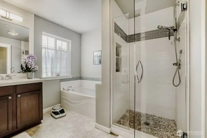 A remodeled walk-in shower has new frameless glass doors, stone tile floor and decorative tile surround with new fixtures.