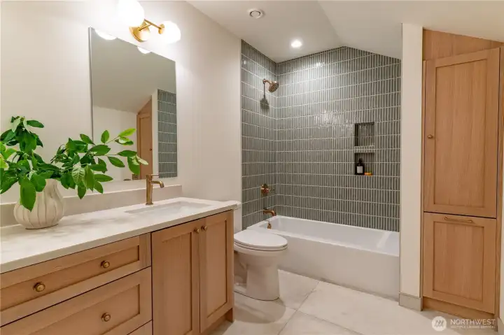 Hall bath with pencil tile surround, white oak vanity, linen tower, and brushed brass fixtures. Heated large format tile floors.