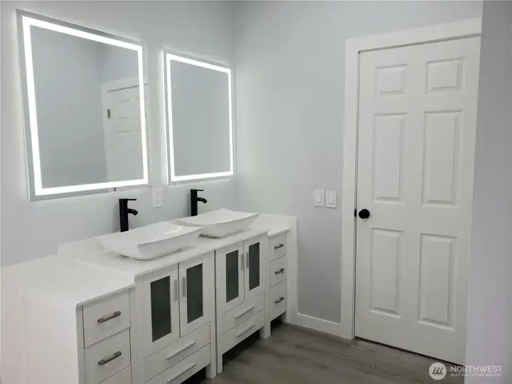 A Full View of the Primary Master Bath Room Showing New Double Sink Vanity, Sinks and Faucet, Two Lighted Mirrors, New Toilet, Paint, Trim and Vinyl Plank Flooring.