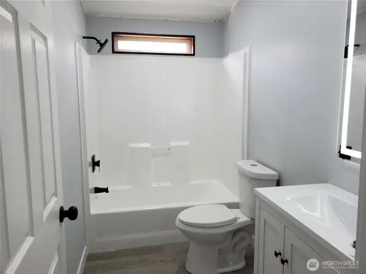 A View of the Main Guest Bath Room Showing New Vanity, Sink and Faucet, a Lighted Mirror, New Toilet, Tub Shower. Paint, Trim and Vinyl Plank Flooring.