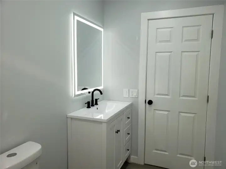 A View of the Main Guest Bath Room Showing New Vanity, Sink and Faucet, a Lighted Mirror, New Toilet, Paint, Trim and Vinyl Plank Flooring.