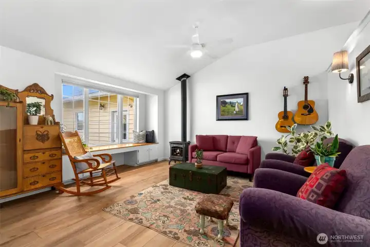 Vaulted-ceiling living room with free-standing propane stove, wood-look tile flooring, and natural light from oversized windows.