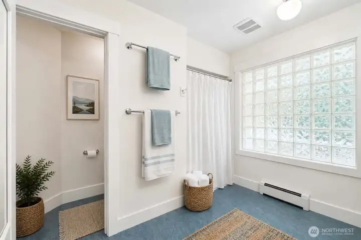virtually staged. Spacious bathroom featuring an easy step-in shower, private toilet room, dual pedestal sinks, and a large glass block window that fills the space with natural light while maintaining privacy.