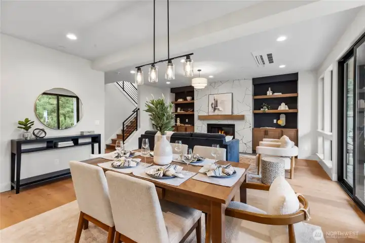 The dining area is bathed in natural light from expansive windows, highlighting the warm tones of the hardwood flooring. A striking multi-bulb light fixture overhead adds a touch of modern elegance and visual interest to the space.
