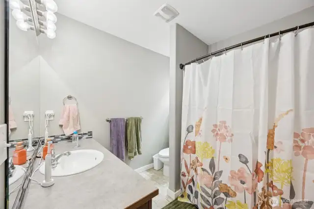 Functional meets thoughtful design in this main bathroom. With vinyl floors that are easy to maintain, a tub and shower combo for versatility, and a long vanity with a spacious mirror, it’s a bathroom designed for both comfort and convenience. The toilet is tucked behind a privacy wall, keeping things neat and tidy, while a window above it provides natural light and ventilation. It’s all about the little details that elevate everyday life.