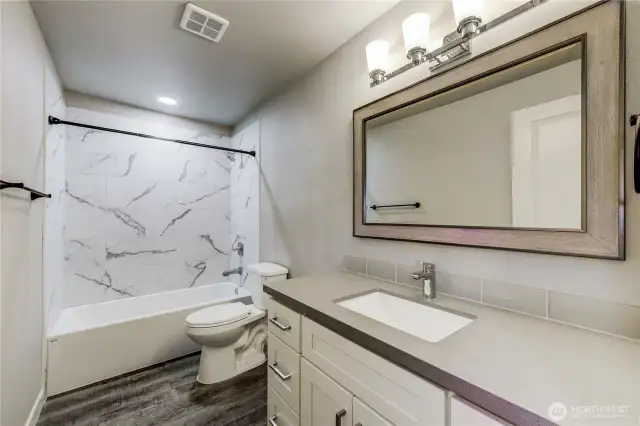 Stylish and updated main bathroom featuring a quartz vanity with sleek modern fixtures, a framed mirror, and elegant lighting. The shower/tub combo is surrounded by striking large-format tile with a marble look,
