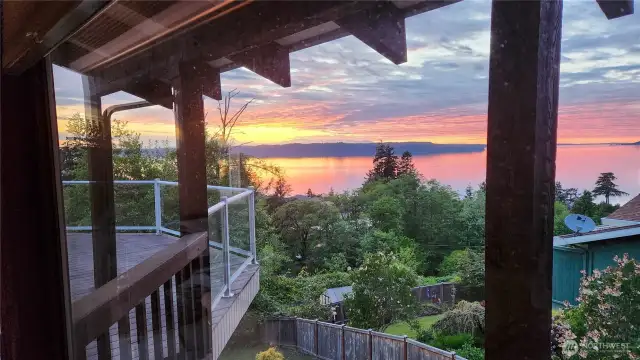 Breathtaking sunset views from the comfort of your own home. This stunning vantage point captures the vibrant colors over Puget Sound, framed by the deck’s elegant glass railing and natural surroundings.