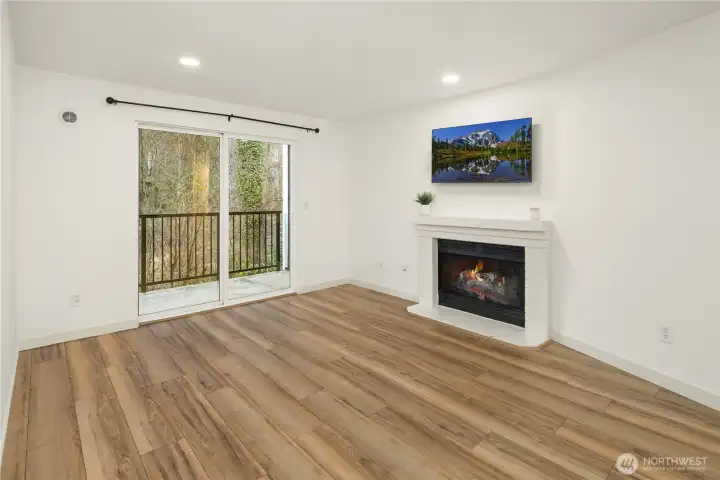 Living area with LVP flooring, TV mount above cozy, wood burning fireplace, and access to balcony with lush greens for peace and quiet.