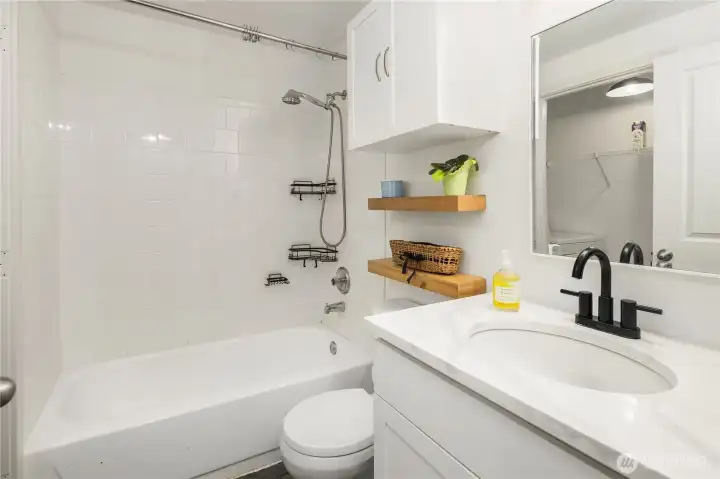 Bathroom with quartz countertops and black durable matte finish faucets. Plenty of storage space in cabinets and wooden, thick shelves. Bath tub with shower products holder in it. Bathroom connected with laundry room.