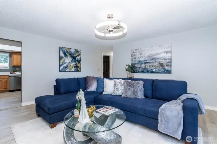 The main living area opens up into a spacious, comfortable setting centered around a classic fireplace that acts as the room’s focal point. The white brick surround adds texture and character without feeling dated, blending nicely with the updated flooring and fresh paint tones. Large windows bring in soft natural light, helping the room feel bright without being harsh. The layout easily accommodates a full sectional while still leaving breathing room, making it ideal for both entertaining and everyday lounging. It’s a practical, livable space that balances warmth and openness.