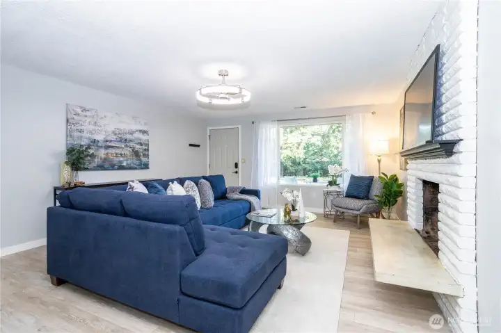 This angle captures the living room from the opposite side, looking out toward the front window and showcasing how much natural light fills the space. The large window acts as a secondary focal point to the fireplace wall, balancing the room with brightness during the day while keeping the atmosphere warm and inviting. From this vantage point, you can see how the layout comfortably accommodates a full sectional while still allowing open walkways and flexible furniture placement. The updated flooring runs seamlessly throughout, reinforcing the home’s refreshed feel. Sightlines toward the entry and adjacent spaces keep everything connected without feeling overly open. It’s a practical, comfortable living area designed for everyday living — whether relaxing, hosting or simply enjoying the natural light that pours in from the front of the home.