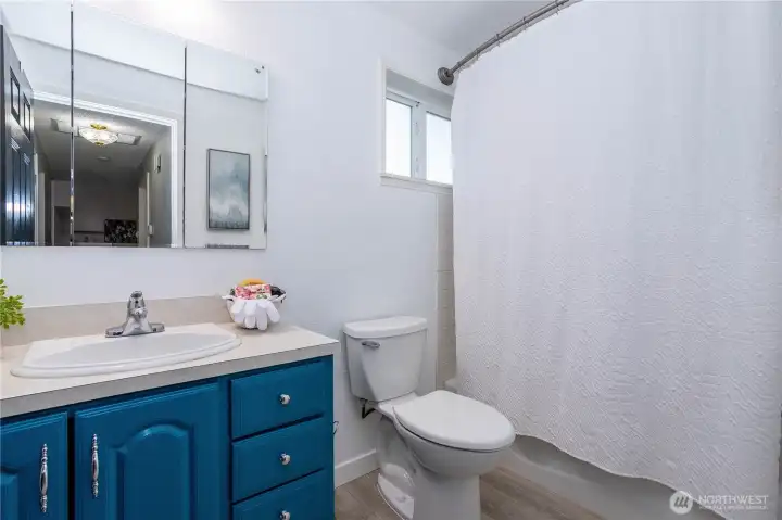 This full bathroom presents clean, functional comfort with a layout designed for everyday ease. The tub and shower combo is wrapped with a full surround and curtain setup, making it practical for both quick mornings and relaxed evening routines. A vanity with storage sits opposite, offering counter space for daily essentials while keeping everything organized. The mirror and lighting keep the room feeling bright, and the neutral finishes make it move-in ready without requiring updates. A small privacy window allows natural light to filter in while maintaining seclusion. Overall, it’s a straightforward, well-kept hall bath positioned conveniently to serve bedrooms and guests alike — practical, clean and ready for daily use.