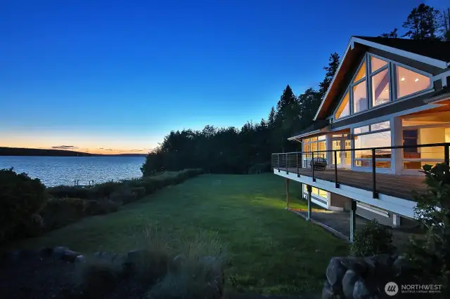 Twilight settles over the expansive backyard as the home glows warmly above the Sound—offering peaceful ambiance, wide-open views, and a stunning connection to the water beyond.