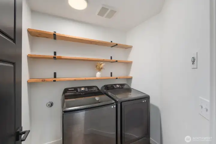 Dedicated laundry room with side-by-side washer and dryer and open shelving above for easy organization of household essentials. A functional utility space designed to keep everyday tasks simple and efficient.