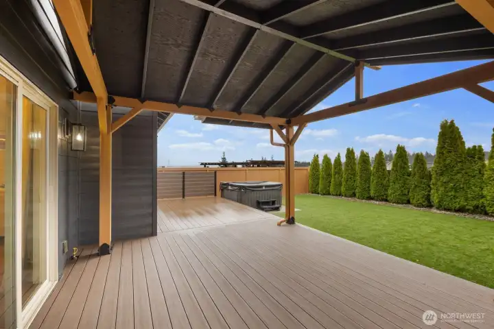 Covered outdoor living area featuring a spacious deck with exposed beam structure overhead, creating a comfortable space for year-round outdoor relaxation and entertaining. The open layout connects directly to the backyard and hot tub area for an inviting outdoor retreat.