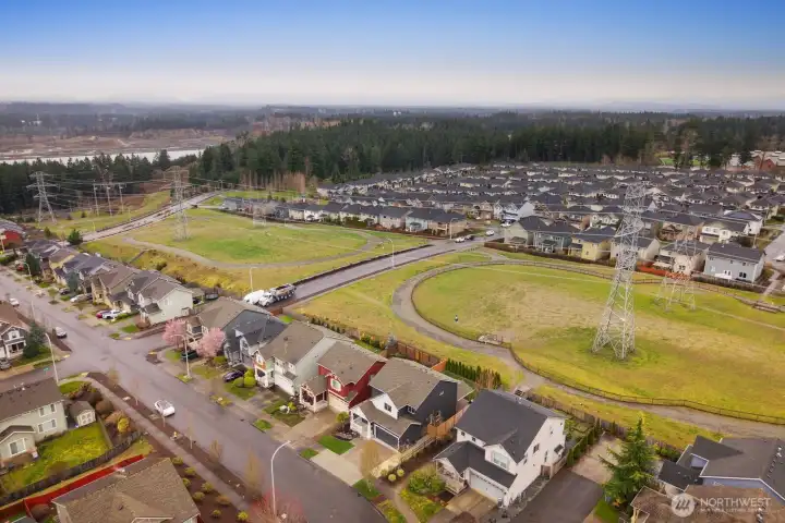 Expanded aerial view capturing the surrounding homes, open fields, and nearby forested landscape. The elevated perspective showcases the balance of neighborhood living and natural surroundings.