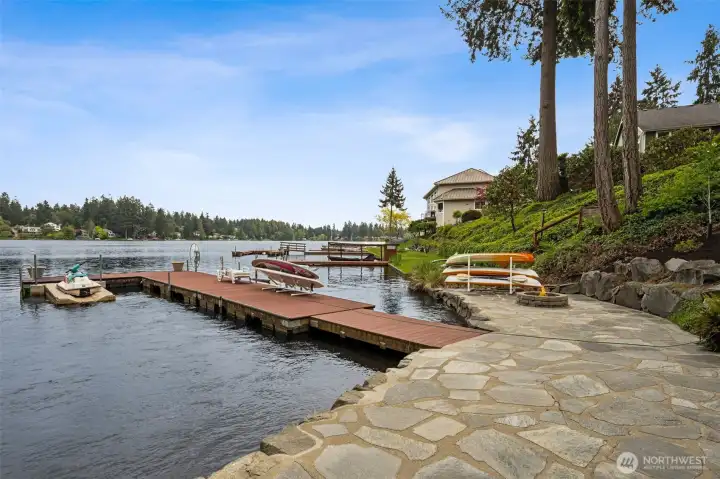 This waterfront hardscaping is absolutely beautiful and completely transforms the shoreline into usable outdoor living space instead of just “yard that happens to touch water.” The wide flagstone-style bulkhead walkway gives you room to set up chairs, lounge seating or front-row sunset viewing spots while still keeping easy access along the water’s edge. And the practical details here are fantastic too. Kayak storage and paddleboard stands sit right near the shoreline for quick grab-and-go lake access, while the nearby outlet and water spigot make cleanup, charging and entertaining dramatically easier. The stonework itself feels substantial and thoughtfully done, creating this polished terraced connection between the lawn, patio and lake that looks incredible while still feeling relaxed and natural within the waterfront setting.