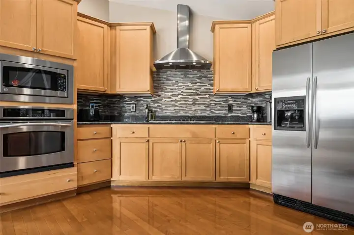 The mix of black granite counters, light maple cabinetry and that gorgeous glass tile backsplash layered in soft creams, grays and warm browns creates so much richness and depth without making the kitchen feel dark or heavy. It is warm, polished and timeless in a way that still feels current years later. The layout works beautifully too, with the wall oven built directly into the cabinetry and a brand-new microwave installed above it keeping everything streamlined and easy to use. Meanwhile the gas cooktop sits below that bold stainless vent hood looking ready for somebody to dramatically sauté something while pretending they are on a cooking show.