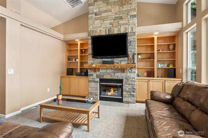 The dramatic floor-to-ceiling stone fireplace already commands attention, but then your eye starts catching all the details surrounding it, the substantial live-edge mantel that actually has presence instead of looking like a decorative shelf from aisle seven, the illuminated built-ins, the shaker cabinetry below and the soft oatmeal carpeting grounding the entire vaulted space in warmth. Even the paint palette is working overtime here in the best possible way. The deeper warm taupe tone rising above the architectural beams adds dimension and contrast, while the softer fawn walls below keep the room feeling bright and inviting.