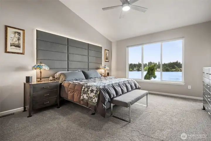 Main-floor living means this home can comfortably evolve with you for years to come, and the bedroom itself feels calm, spacious and wonderfully connected to the lake. The vaulted ceiling adds volume overhead while that massive three-panel picture window practically turns Long Lake into the headboard feature wall. It is the kind of view people immediately walk toward the second they enter the room. The brand-new carpeting in that rich medium brown tone keeps everything grounded and cozy, while the ceiling fan adds year-round comfort without disrupting the clean look of the space, and the electric shades aid to darken the room during any day sleeping.