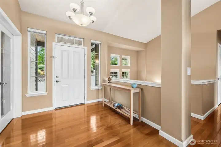 This foyer walks the line between grand and welcoming beautifully. Natural light pours in from every direction thanks to tall accent windows flanking the front door, plus another transom window above that keeps the entire entry glowing from the second you walk in. The cherry-toned wood floors bring richness and warmth underfoot, while the soft fawn wall color paired with crisp white millwork keeps everything feeling timeless instead of trendy. And then your eye starts traveling through the architectural details. The half-wall staircase separation keeps the entry wonderfully open, while that cluster of four windows climbing above the staircase adds such a cool visual rhythm and dimension to the space.