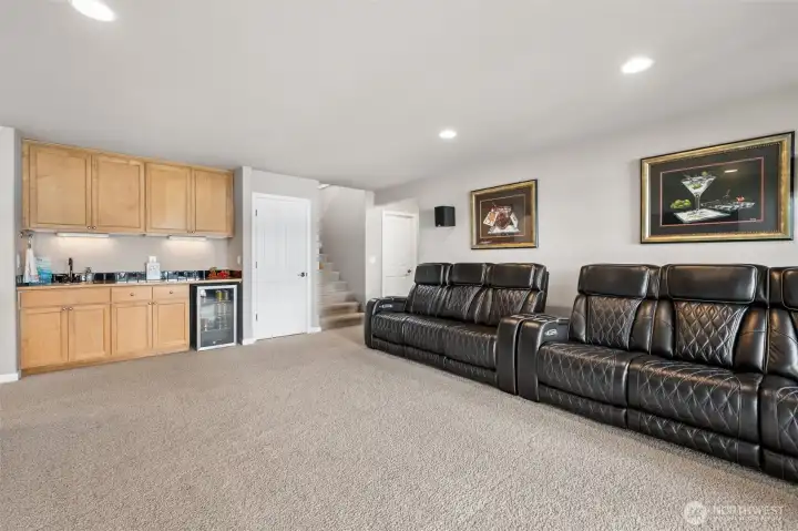 Six full recliners fit comfortably along one wall and the space still feels open, which tells you everything you need to know about how well this lower level handles gatherings. The beverage bar setup is fantastic too, complete with sink, beverage fridge and an impressive amount of maple cabinetry above and below for storing everything from glassware to board games to emergency lake-house snacks. Then there is the giant under-stair storage closet beside the beverage fridge, affectionately giving strong Harry Potter closet energy except significantly more useful and far less emotionally traumatic. Holiday décor, extra life jackets, Costco overflow, this thing swallows storage needs whole. And the layout continues to shine, with the hallway door on the right leading toward the opposite downstairs wing containing another bedroom, bathroom and huge storage room, giving the lower level incredible flexibility and separation for guests or multigenerational living.