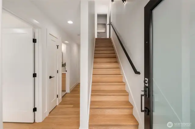A welcoming foyer leads past main-floor bedrooms and bath, with warm wood floors and a clean modern staircase drawing you up to the open-concept living and kitchen spaces above.