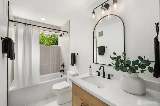 Fresh, modern style in the main floor bathroom with a frameless arched mirror, stylish vanity lights, vertical tile surround, and a deep soaking tub paired with a shower, set beneath a bright window.
