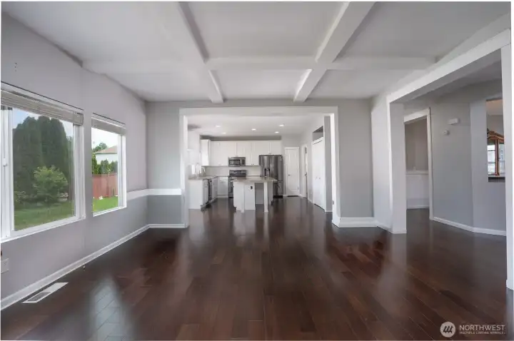Bright and spacious open-concept living area featuring rich hardwood flooring, large windows for abundant natural light, and elegant coffered ceilings that add architectural character and style. The seamless layout flows directly into the modern kitchen, complete with white cabinetry, stainless steel appliances, granite countertops, and a center island perfect for gathering and entertaining. Neutral tones and clean finishes throughout create a warm, inviting space ready to make your own.